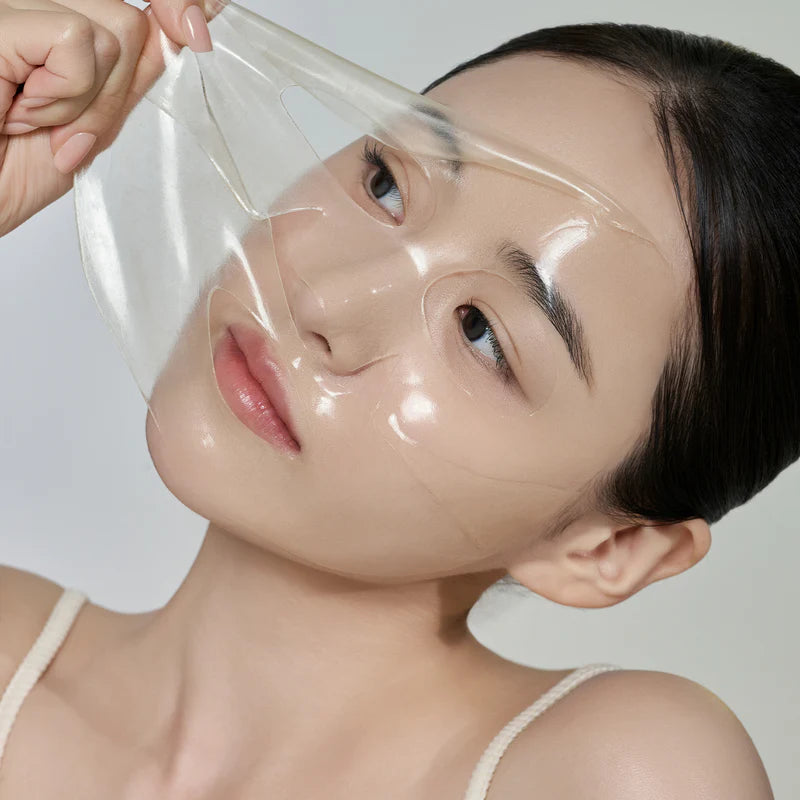 Uma modelo coreana retira delicadamente uma máscara facial transparente, revelando pele lisa por baixo. A sua expressão é serena e o fundo está suavemente iluminado. KBEauty. Cosmetica Coreana