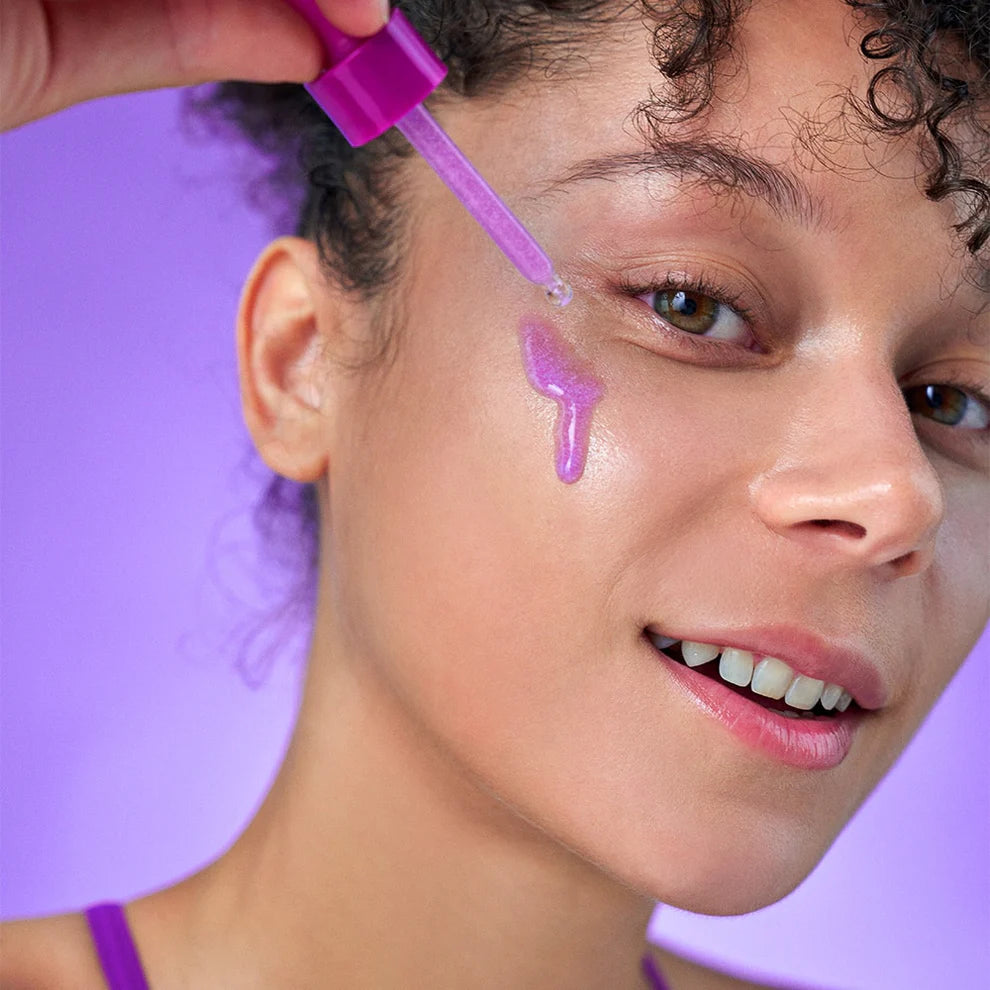 Uma mulher sorri enquanto aplica um sérum roxo Bakuchiol Plumping Serum da eqqualberry na bochecha com um conta-gotas. O fundo é um tom roxo suave, que complementa a sua expressão alegre.