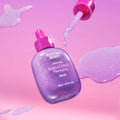 Um frasco de vidro de Bakuchiol Plumping Serum roxo, com uma tampa conta-gotas cor-de-rosa, flutua sobre um fundo rosa suave. Uma gota de sérum está suspensa no ar.