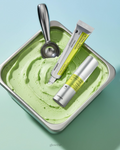 Um recipiente de metal cheio de sorvete cremoso verde é coberto com uma colher de sorvete prateada e dois cosmeticos coreanos  produtos para cuidados com a pele. A cena é leve e refrescante.