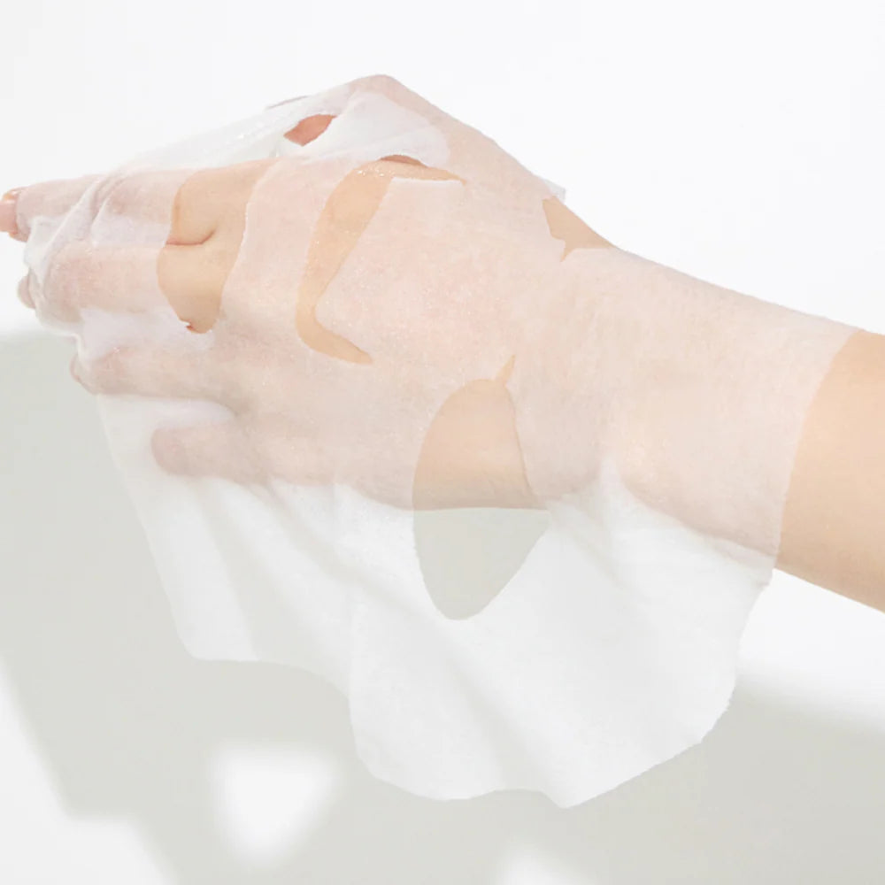 Uma mão segura uma máscara facial branca e semi-transparente - Real Shea Butter Essence Mask - sobre um fundo neutro e claro. A máscara cobre os dedos, transmitindo a sensação de um ritual de cuidados de pele.