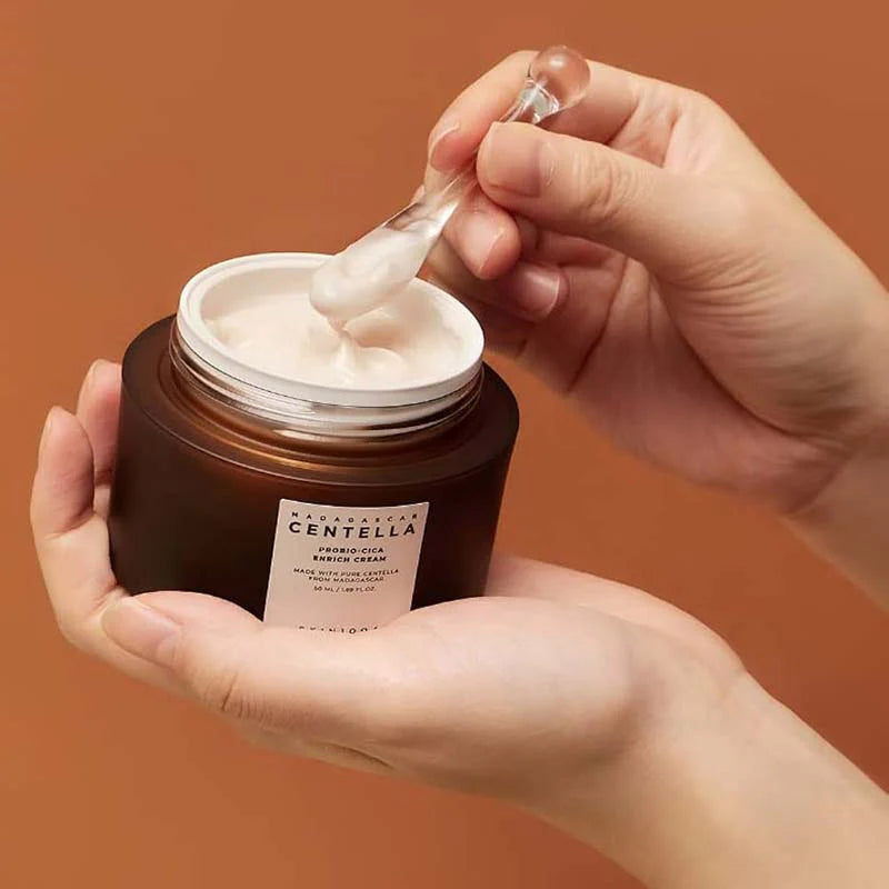 Mãos seguram o Madagascar Centella Probio-Cica Enrich Cream da SKIN1004 um frasco castanho de creme de cuidados de pele sobre um fundo castanho quente. Uma espátula com creme suave é mergulhada no frasco, transmitindo uma sensação de luxo.