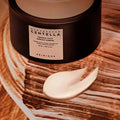Recipiente escuro de Centella Probio-Cica Cream da SKIN1004 sobre uma superfície texturada em tons terrosos. Uma pequena espiral de creme está colocada à frente, transmitindo uma sensação de luxo.
