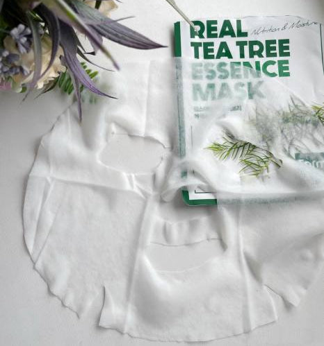 Máscara facial de essência de tea tree colocada sobre uma superfície branca, ao lado da sua embalagem, com flores próximas, criando uma estética fresca e relaxante.