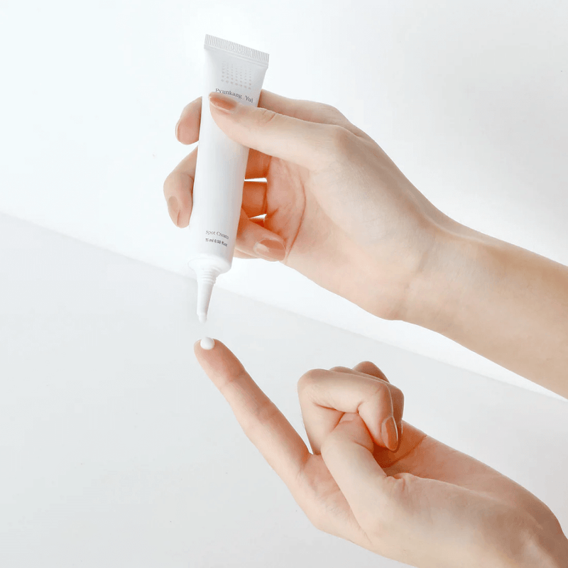 Close-up de mãos a aplicar uma pequena quantidade de creme branco Acne spot cream da marca coreana pyunkang yul - de um tubo na ponta do dedo. O ambiente é minimalista e luminoso, transmitindo uma sensação de limpeza e calma. Cosmética.