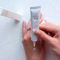 Uma mão a aplicar o Acne Spot Cream da marca coreana Pyunkang Yul nas costas de outra mão. Um tubo branco é espremido, com uma caixa ao fundo. A cena transmite calma e concentração. Cosmetica Coreana em Portugal.