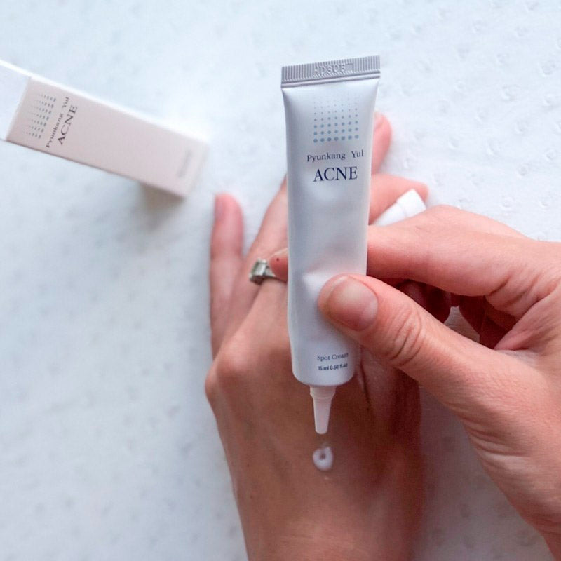 Uma mão a aplicar o Acne Spot Cream da marca coreana Pyunkang Yul nas costas de outra mão. Um tubo branco é espremido, com uma caixa ao fundo. A cena transmite calma e concentração. Cosmetica Coreana em Portugal.