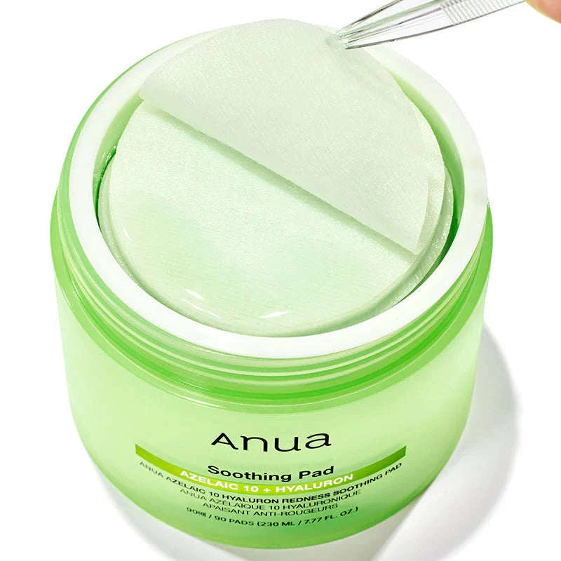 Um frasco verde com a inscrição “Anua Soothing Pad” contém discos circulares embebidos em ácido azelaico e hialuron. Uma pinça levanta um dos discos, sugerindo frescura e cuidado. KBeauty.