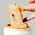 Uma mão mergulha num frasco de Calendula Rice Mochi Cleanser espessa e cremosa, esticando-a para cima. A textura aparenta ser suave e pegajosa, transmitindo uma sensação t