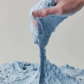 Uma mão estica uma o mochi rice cleanser azul, com pequenos pontos pretos, sobre um fundo simples. A textura aparenta ser suave e pegajosa, transmitindo uma sensação divertida.