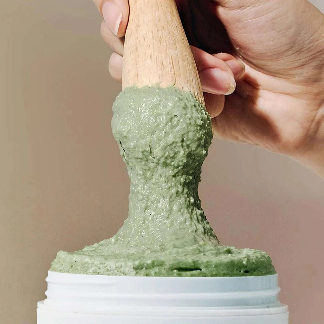 Uma mão segurando uma espátula de madeira mergulha num frasco de FRESH GREEN RICE MOCHI CLEANSER da arencia
. A textura aparenta ser suave e cremosa sobre u