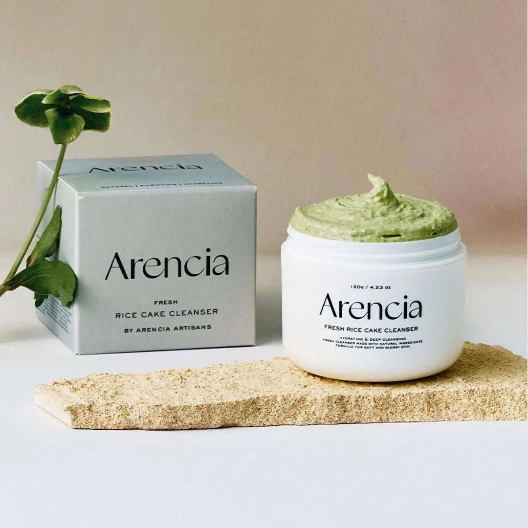 Frasco branco do Arencia Fresh Rice Cake Cleanser com pasta verde sobre uma superfície arenosa, acompanhado pela sua caixa e por uma pequena planta verde, criando uma estética fresca e natural.
