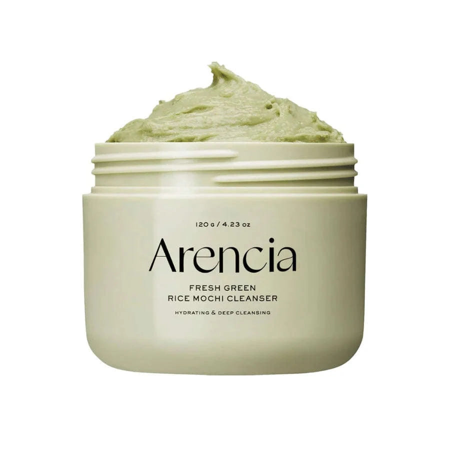 Frasco branco do Arencia Fresh Rice Cake Cleanser com pasta verde sobre uma superfície arenosa, acompanhado pela sua caixa e por uma pequena planta verde, criando uma estética fresca e natural.