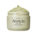 Frasco branco do Arencia Fresh Rice Cake Cleanser com pasta verde sobre uma superfície arenosa, acompanhado pela sua caixa e por uma pequena planta verde, criando uma estética fresca e natural.