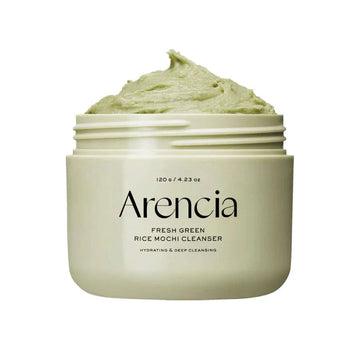 Frasco branco do Arencia Fresh Rice Cake Cleanser com pasta verde sobre uma superfície arenosa, acompanhado pela sua caixa e por uma pequena planta verde, criando uma estética fresca e natural.