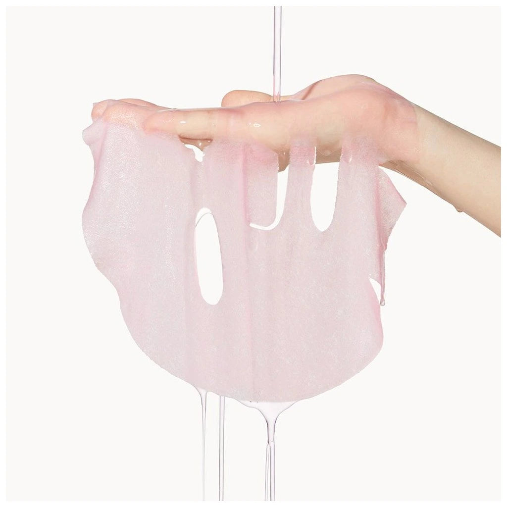 Uma mão segura uma máscara facial rosa translúcida e húmida, pingando água. A textura é delicada e suave, transmitindo uma sensação de frescura e cuidado com a pele.