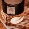 Recipiente escuro de Centella Probio-Cica Cream da SKIN1004 sobre uma superfície texturada em tons terrosos. Uma pequena espiral de creme está colocada à frente, transmitindo uma sensação de luxo.