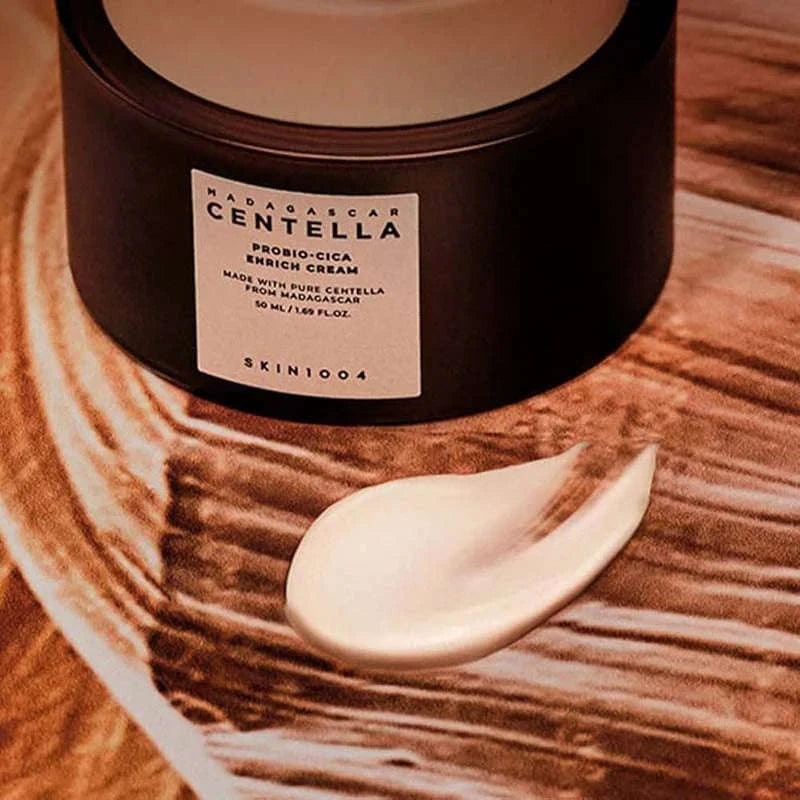 Recipiente escuro de Centella Probio-Cica Cream da SKIN1004 sobre uma superfície texturada em tons terrosos. Uma pequena espiral de creme está colocada à frente, transmitindo uma sensação de luxo.