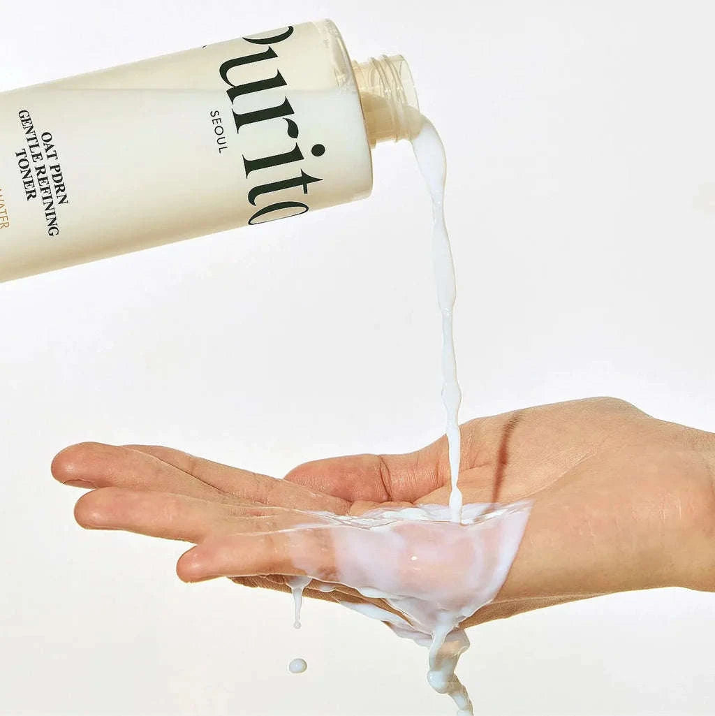 Uma mão verte o tónico Oat PDRN Gentle Refining Toner líquido de textura leitosa de um frasco para a palma da mão. A imagem transmite uma sensação de frescura e limpeza, com uma estética minimalista.