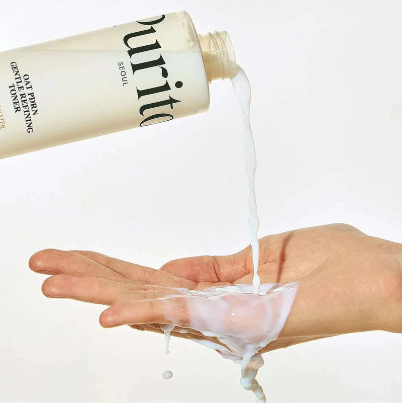 Uma mão verte o tónico Oat PDRN Gentle Refining Toner líquido de textura leitosa de um frasco para a palma da mão. A imagem transmite uma sensação de frescura e limpeza, com uma estética minimalista.