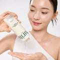Uma mulher com pele luminosa sorri enquanto verte o tónico Oat PDRN Gentle Refining Toner de um frasco para a mão. A imagem transmite uma sensação de frescura e de ritual de cuidados de pele.