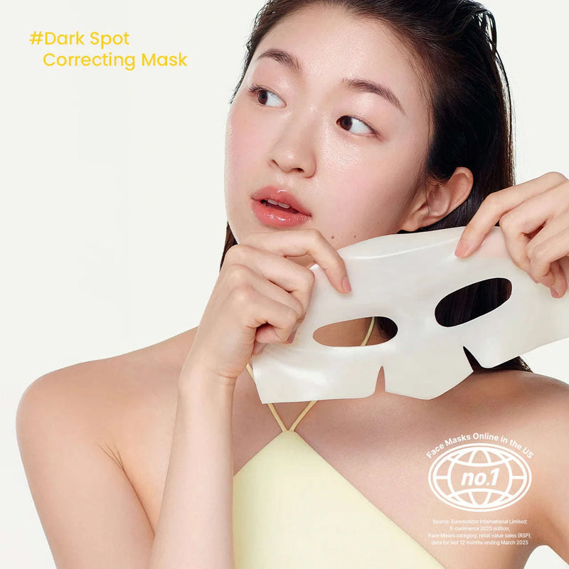Uma mulher com pele radiante está a retirar uma máscara facial branca, sorrindo ligeiramente, sobre um fundo simples. O texto indica: “#Máscara Corretora de Manchas Escuras.” KBeauty. Biodance Mask.