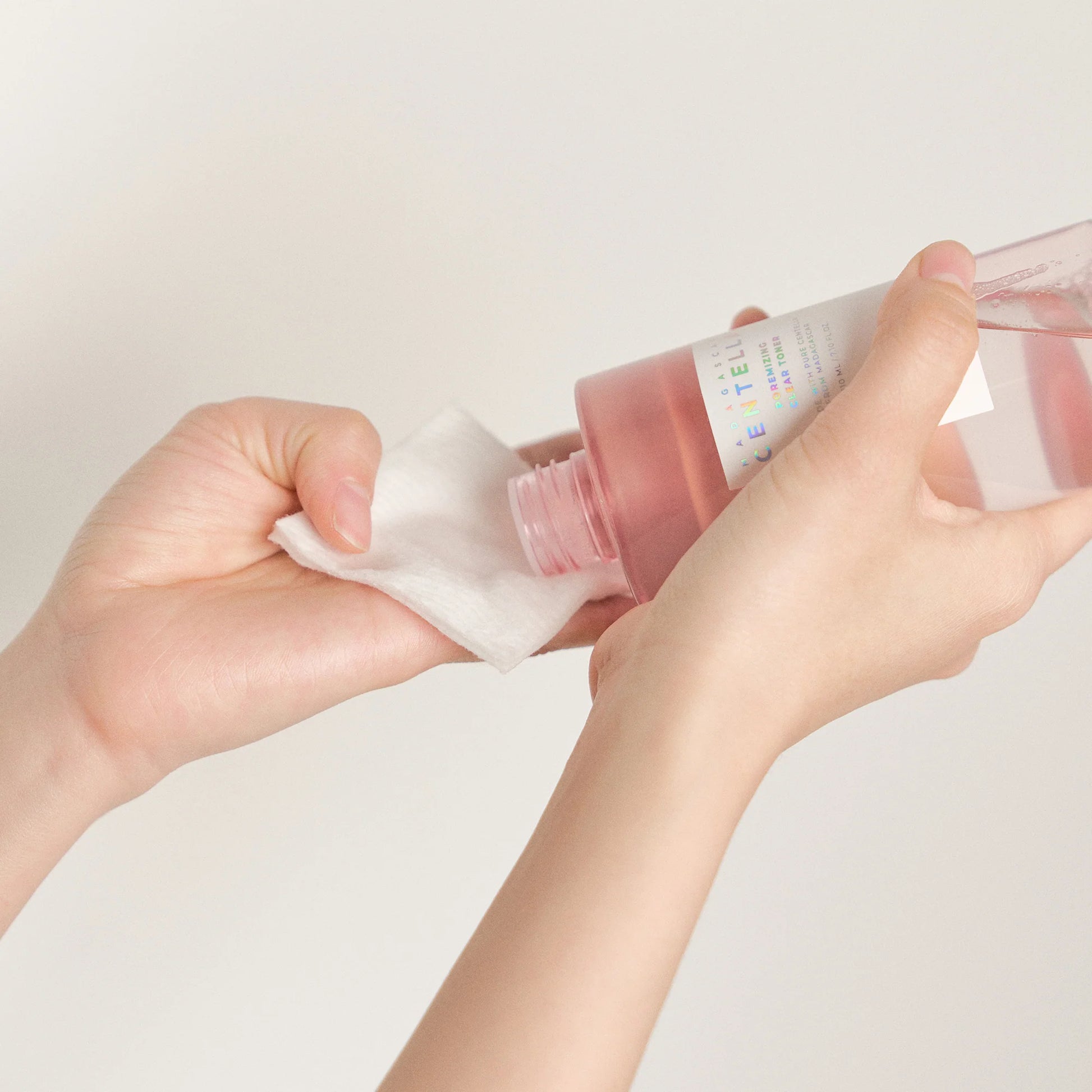 Duas mãos seguram um frasco rosa de 210ml,  de SKIN1004 Madagascar Centella Poremizing Clear Toner, vertendo o tonico facial sobre um disco de algodão branco. O fundo é simples, destacando uma rotina de cuidados de pele.