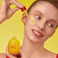 Uma pessoa sorridente aplica sérum Vitamin illuminating Serum vitamínico da EQQUALBERRY na bochecha com um conta-gotas, sobre um fundo amarelo vivo, transmitindo uma sensação de frescura e luminosidade.